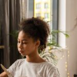 Etiquette - Serious African American girl with unrecognizable mother sitting at wooden table with plate and silverware in hands learning table manners